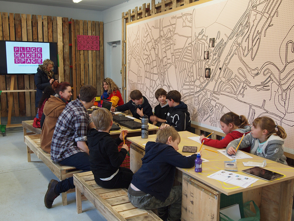 Group of school children sat at a wooden bench, drawing and discussing with large map on wall behind.