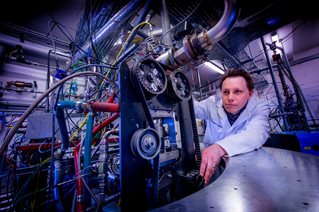 Engineering research in smart transport being conducted on machinery at the University of Brighton Advanced Engineering Centre. Male researcher in lab coat examines belt drive and cogs with surrounding tubes, transparent, red and blue.
