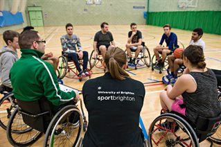 Wheelchair sports players in a circle inside a gym. The back of one person's shirt has the Sport Brighton University of Brighton logo